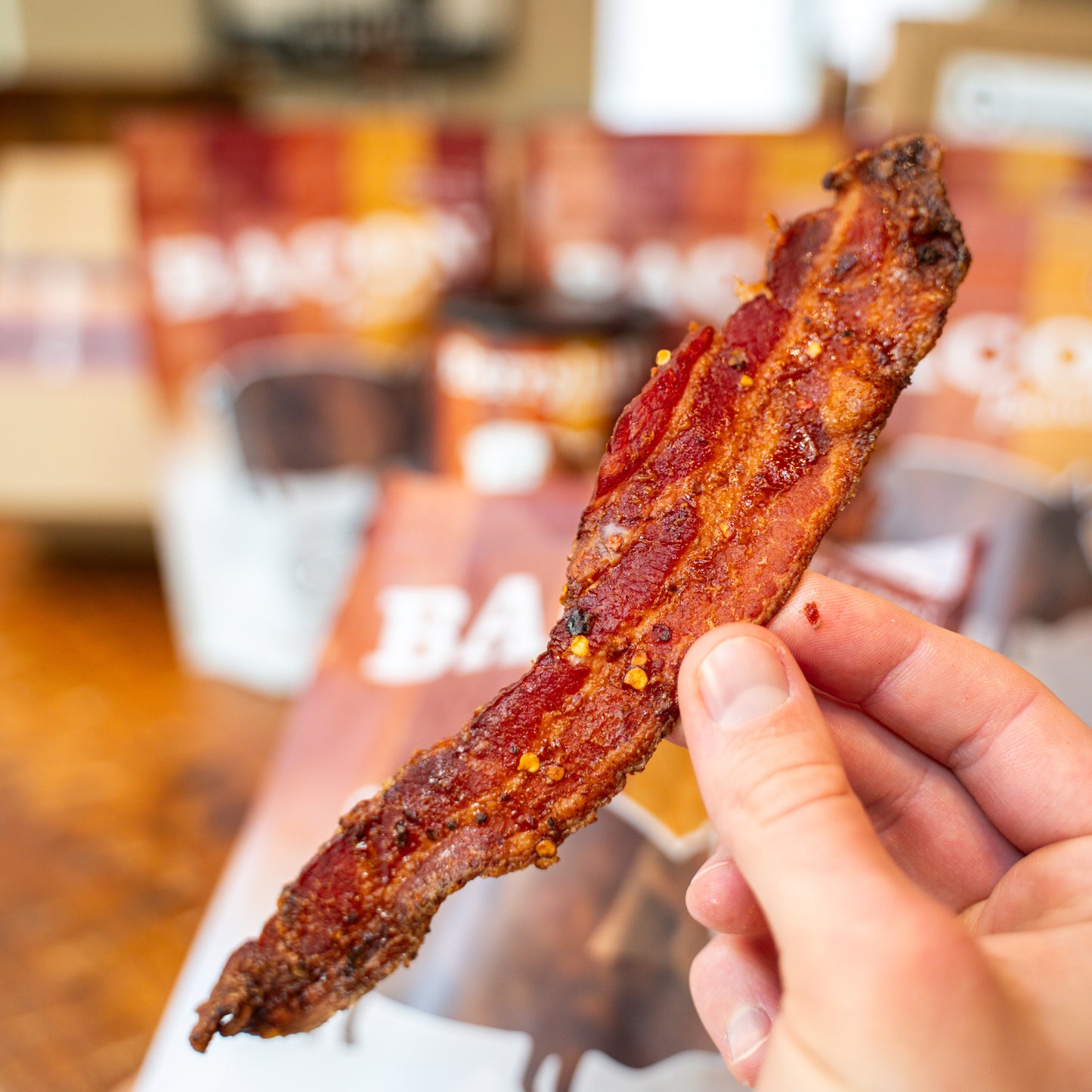 Person holding a slice of bacon with a blurred restaurant interior in the background