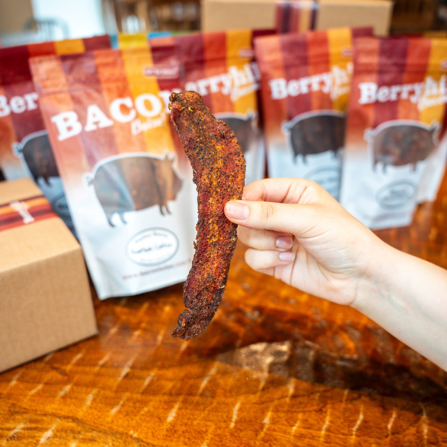 A hand holding a piece of bourbon coffee bacon with packages of Berryhill Bacon products in the background.