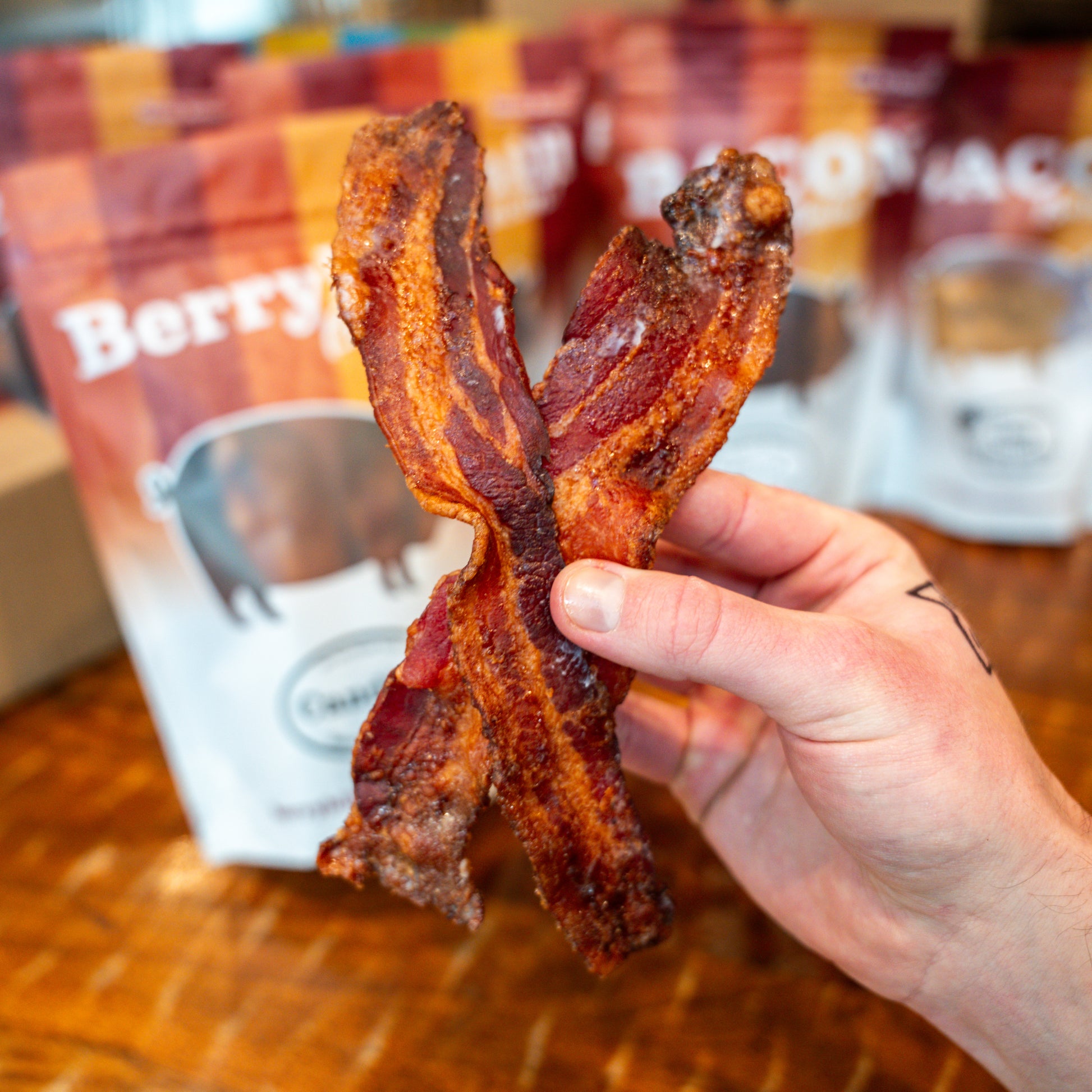 A hand holding a piece of crispy bacon with a package of Berryhill Bacon visible in the background.