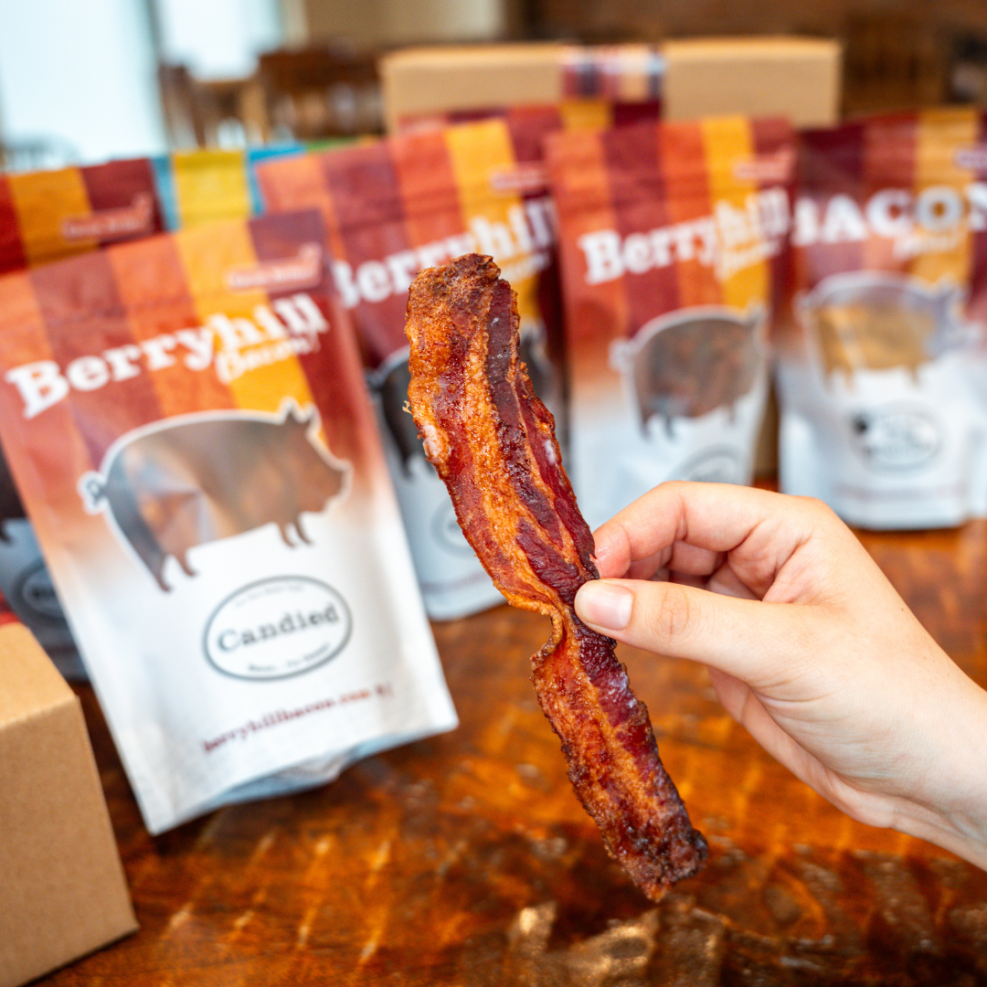 A hand holding a piece of candied bacon with packages of Berryhill Candied Bacon visible in the background.