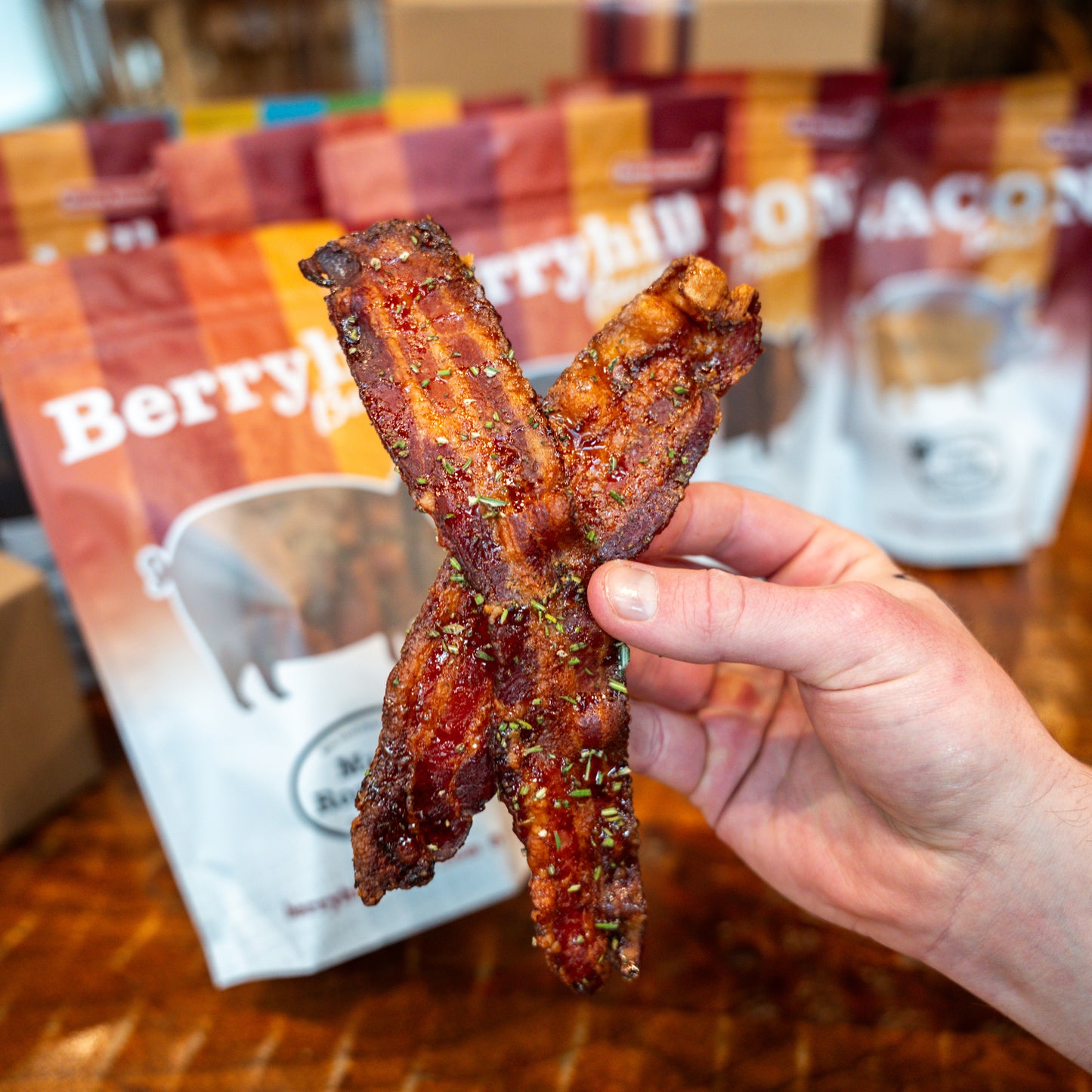 A hand holding a piece of crispy bacon with a package of Berryhill Bacon visible in the background.