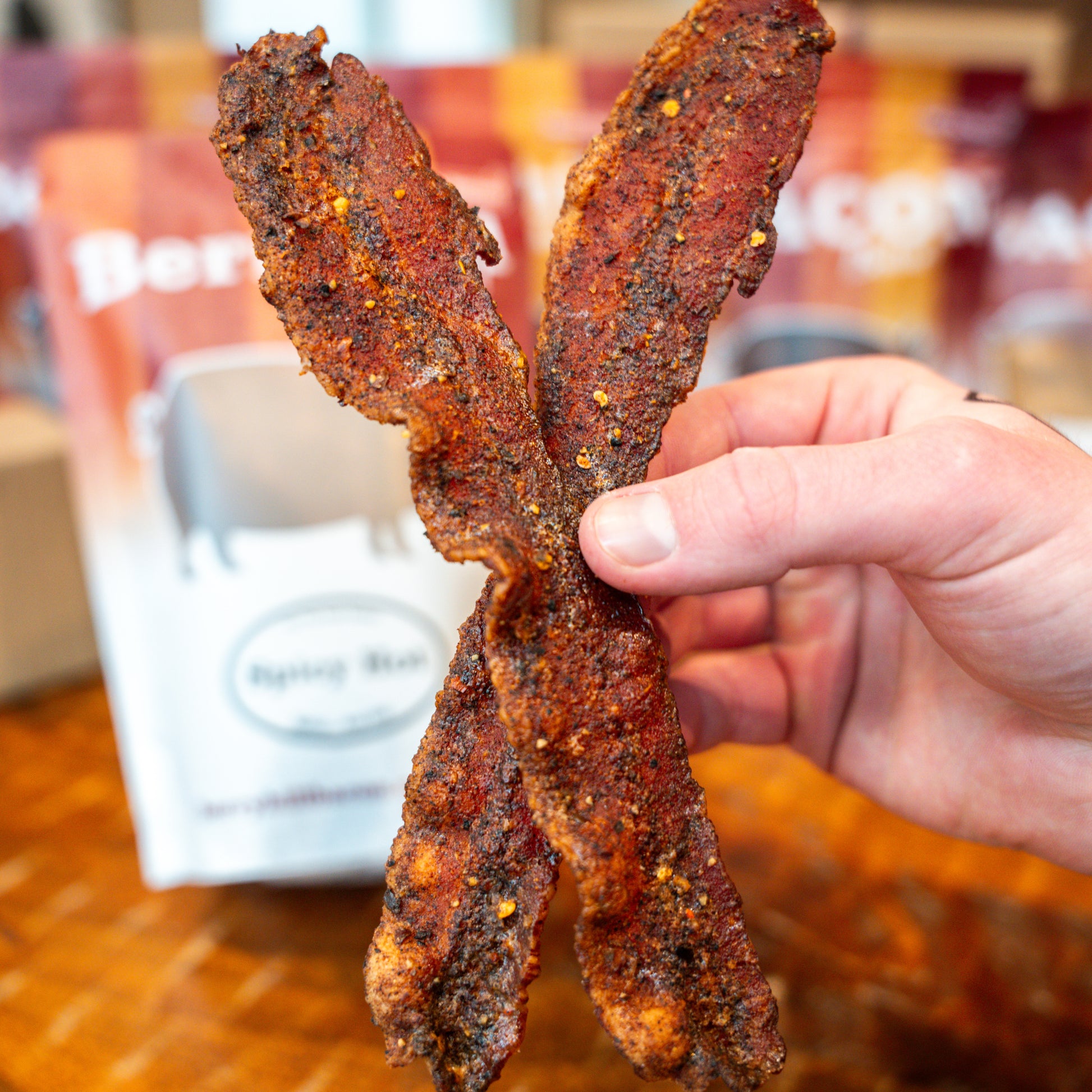 A hand holding a piece of crispy bacon with a package of Berryhill Bacon visible in the background.