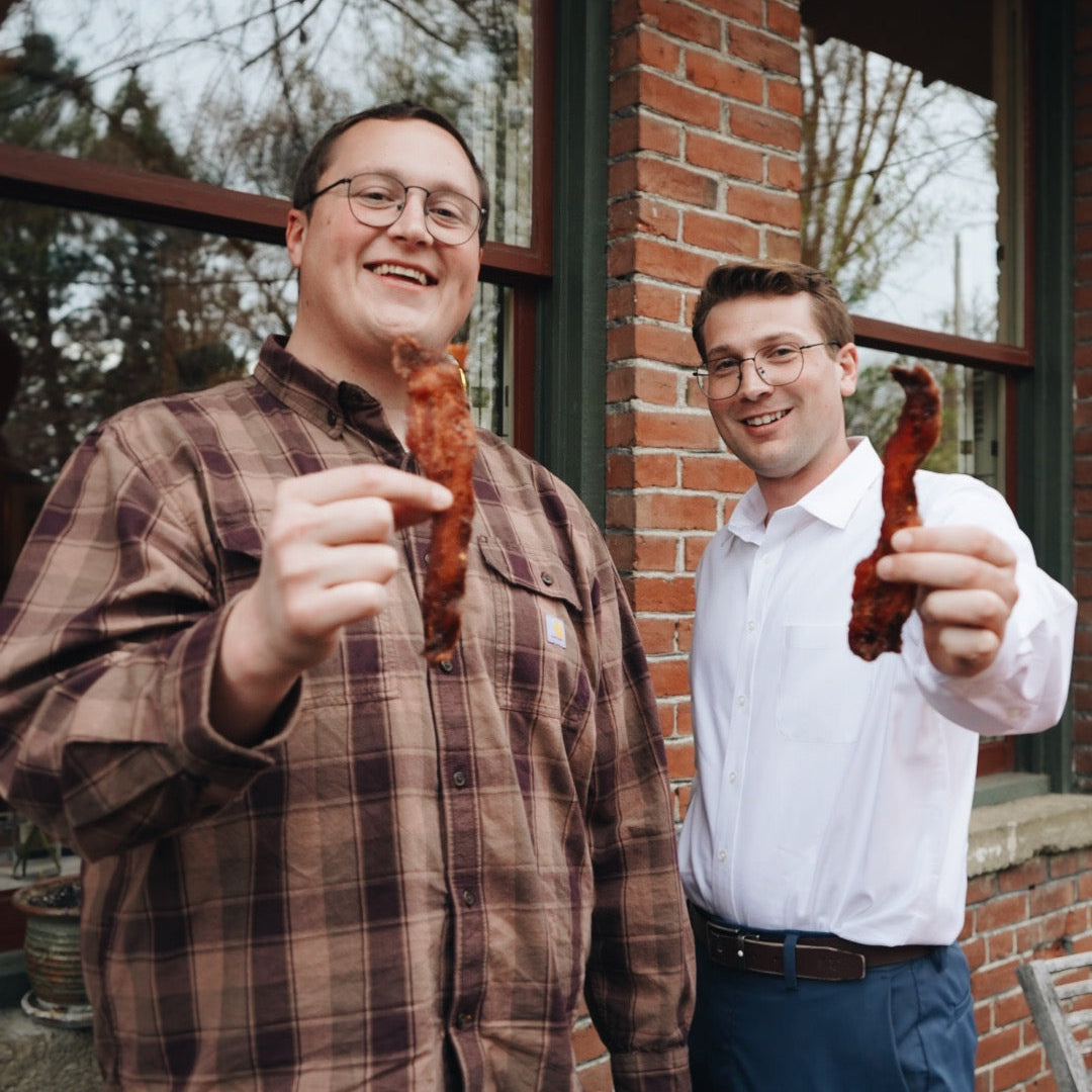 Two men holding pieces of bacon outdoors in front of a brick building.