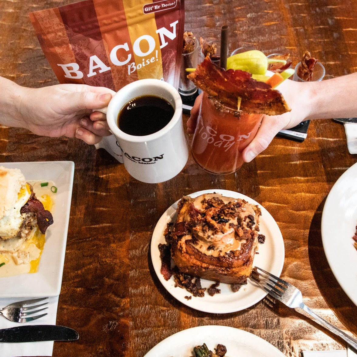 A cup of coffee in a BACON Boise white mug and a Bloody Mary "cheersing" over a wooden table with a bacon cinnamon roll.