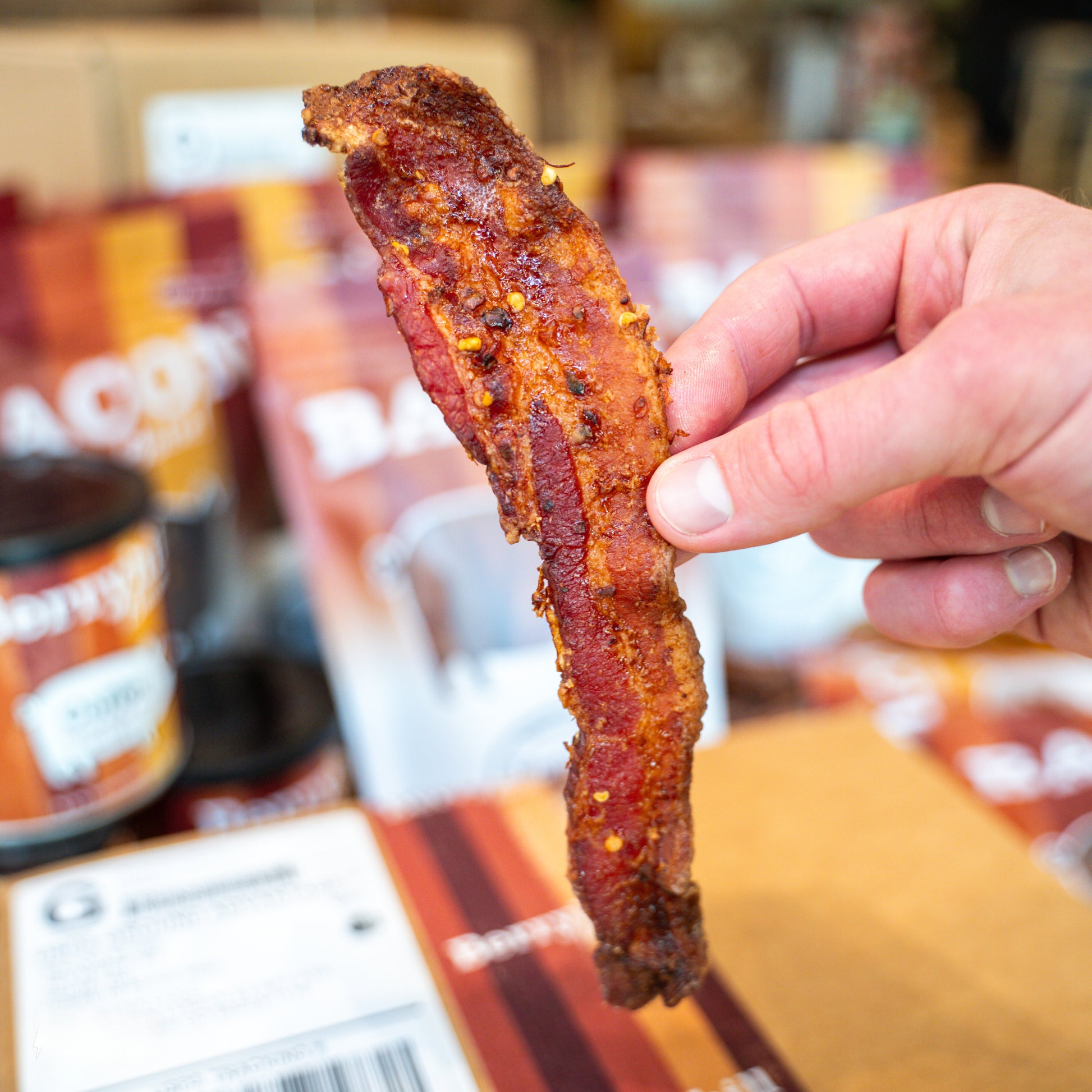 A hand holding a piece of crispy bacon with a package of Berryhill Bacon visible in the background.