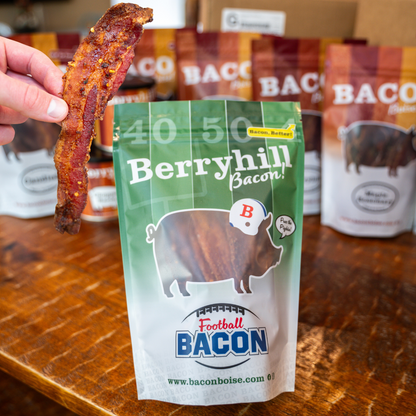 A hand holding a piece of bacon with a package of Berryhill Bacon visible in the foreground. The packaging has branding and the text 'Football Bacon' on it.