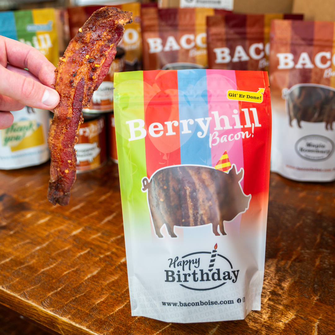 A packaged piece of bacon with a 'Berryhill Bacon' branding next to a box labeled 'BACON Boise'. The bacon package has a 'Happy Birthday' theme with a pig illustration and birthday decorations.