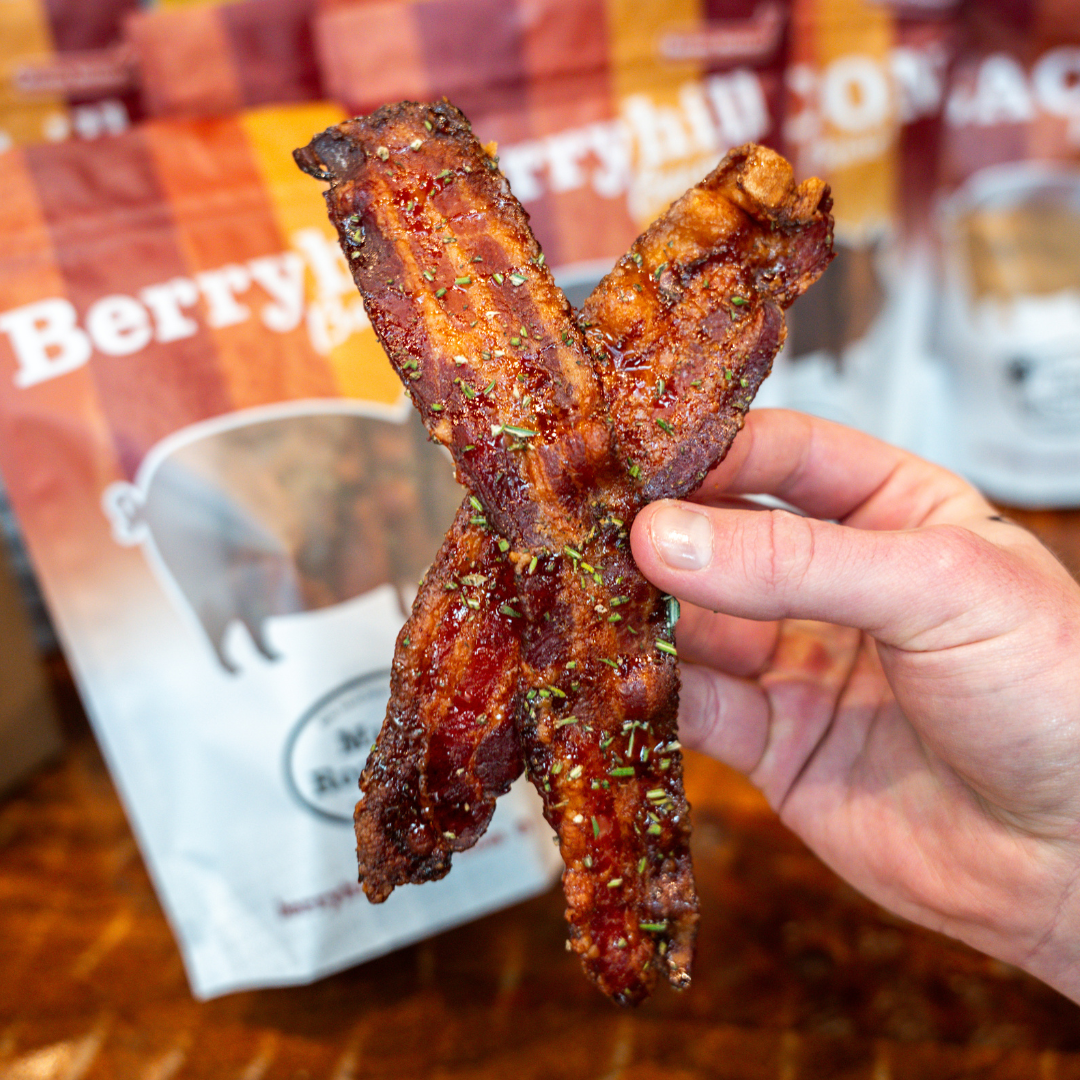 A hand holding a piece of Maple Rosemary Bacon, with a package of Berryhill Bacon visible in the background.