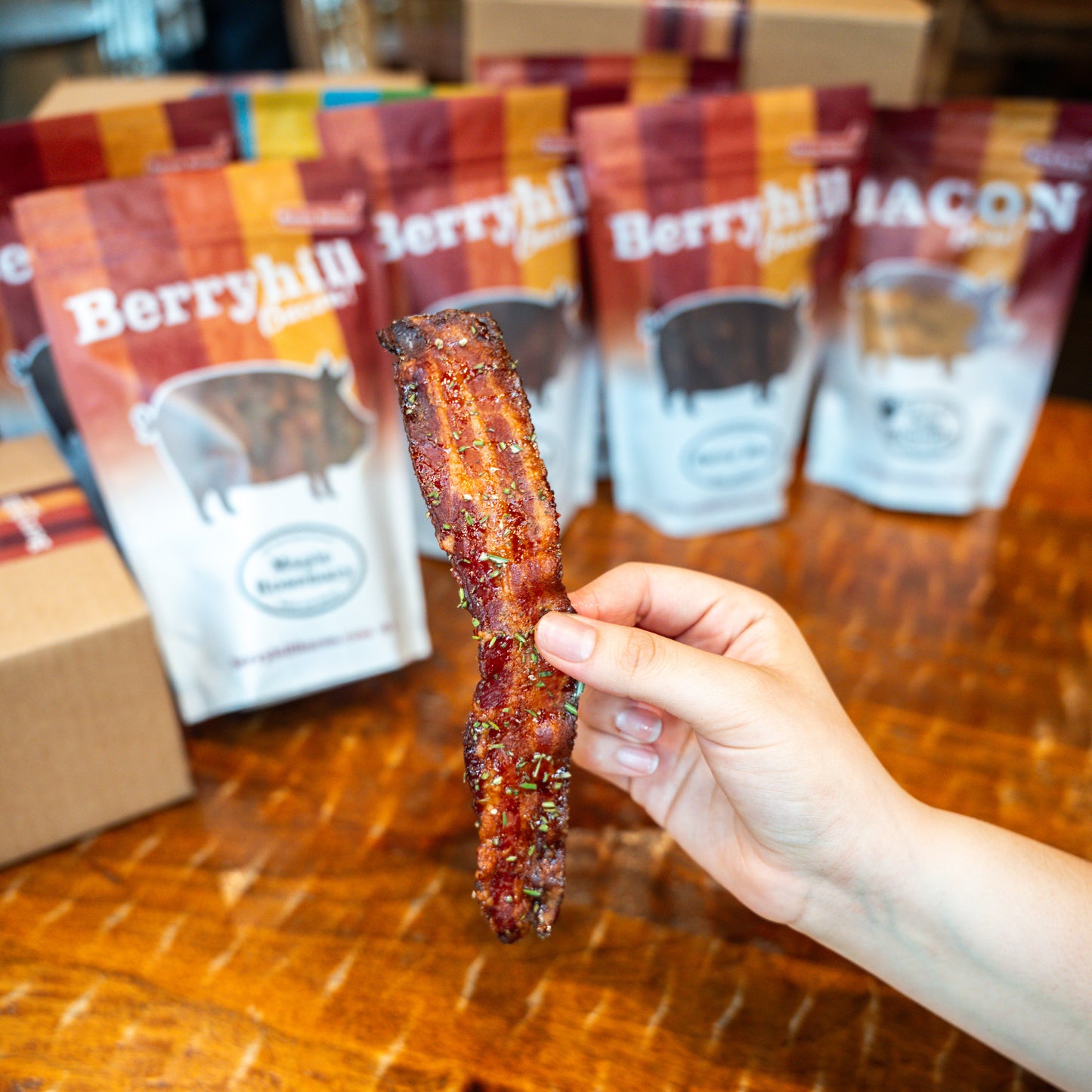 A hand holding a strip of Berryhill Bacon with a bacon bag labeled maple rosemary faded but visible behind, sitting on a wooden table. 
