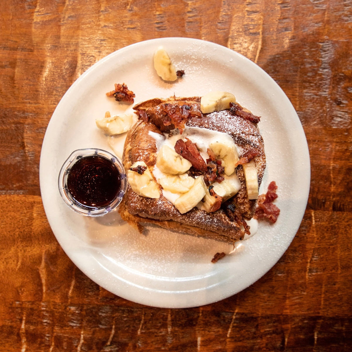 French toast with chopped bacon and banana on a white plate with a small bowl of syrup on a wooden table.