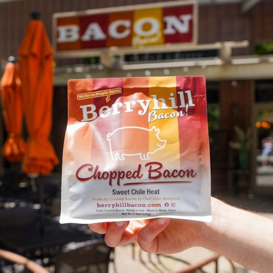 Hand holding a package of Berryhill Bacon chopped bacon in front of a building with 'Bacon' sign.
