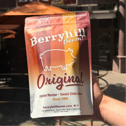 A hand holding a bag of Berryhill Bacon Original in front of an outdoor restaurant setting with umbrellas and a bacon sign in the background.