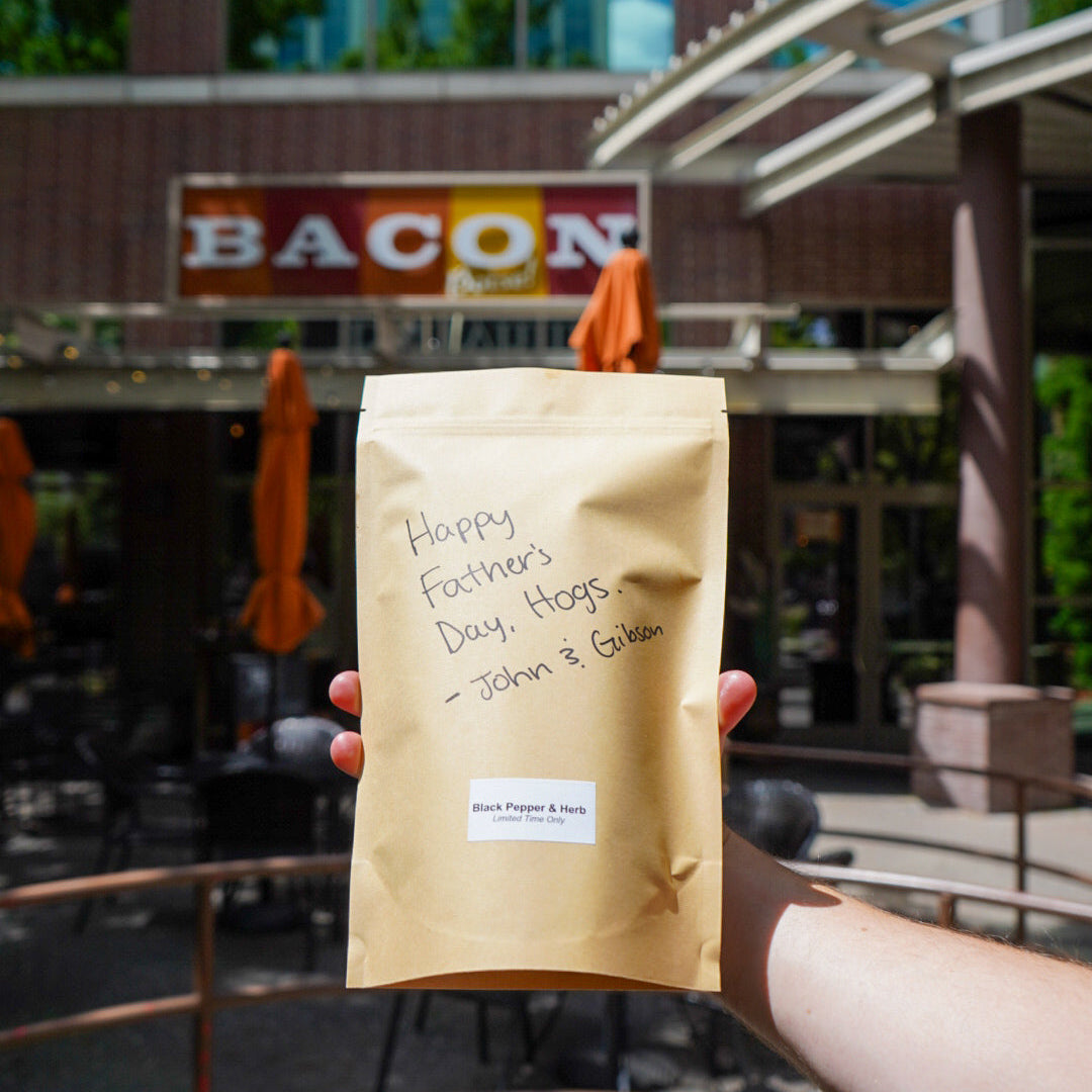 Hand holding a coffee package with 'Happy Father's Day' message in front of a building with a sign that reads 'Bacon'.