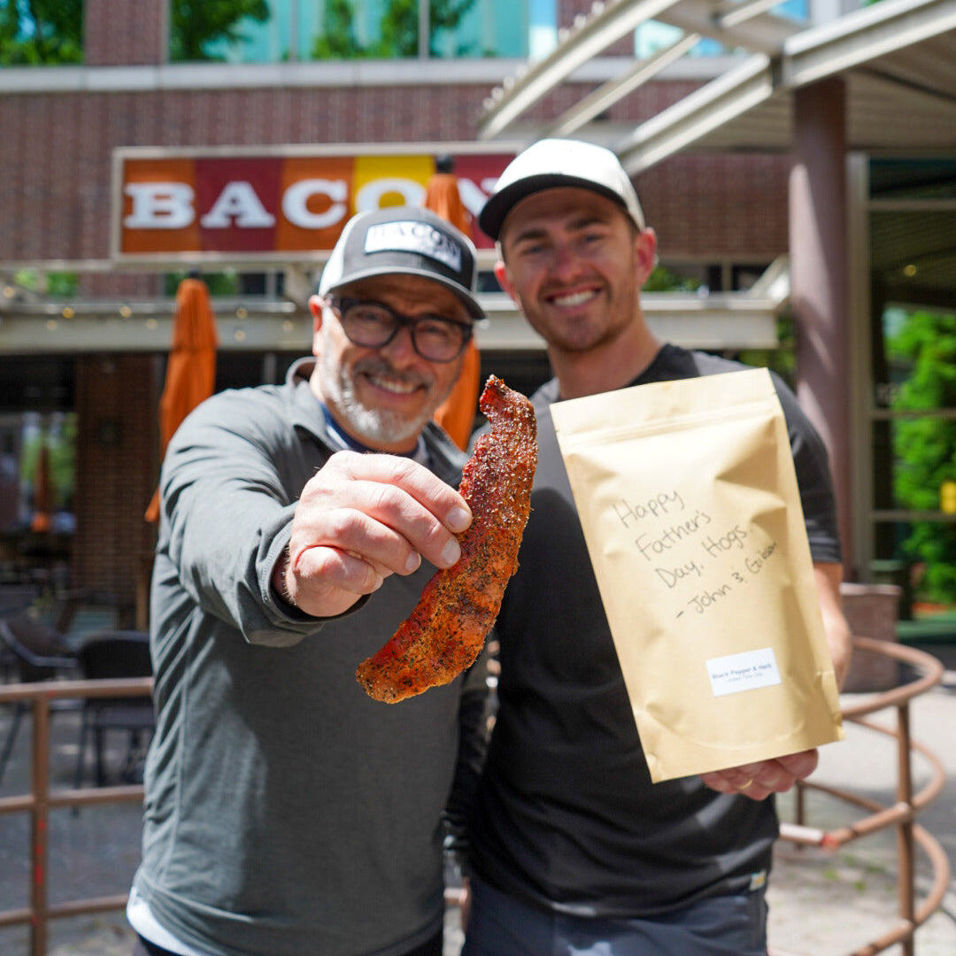 Two men holding a bag of bacon and a piece of bacon outdoors with a building in the background.