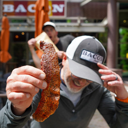 Man holding a large piece of bacon outdoors with a 'BACON' sign in the background.