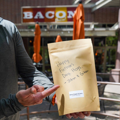 Person holding a bacon package with a message outdoors, with 'Bacon' sign in the background.