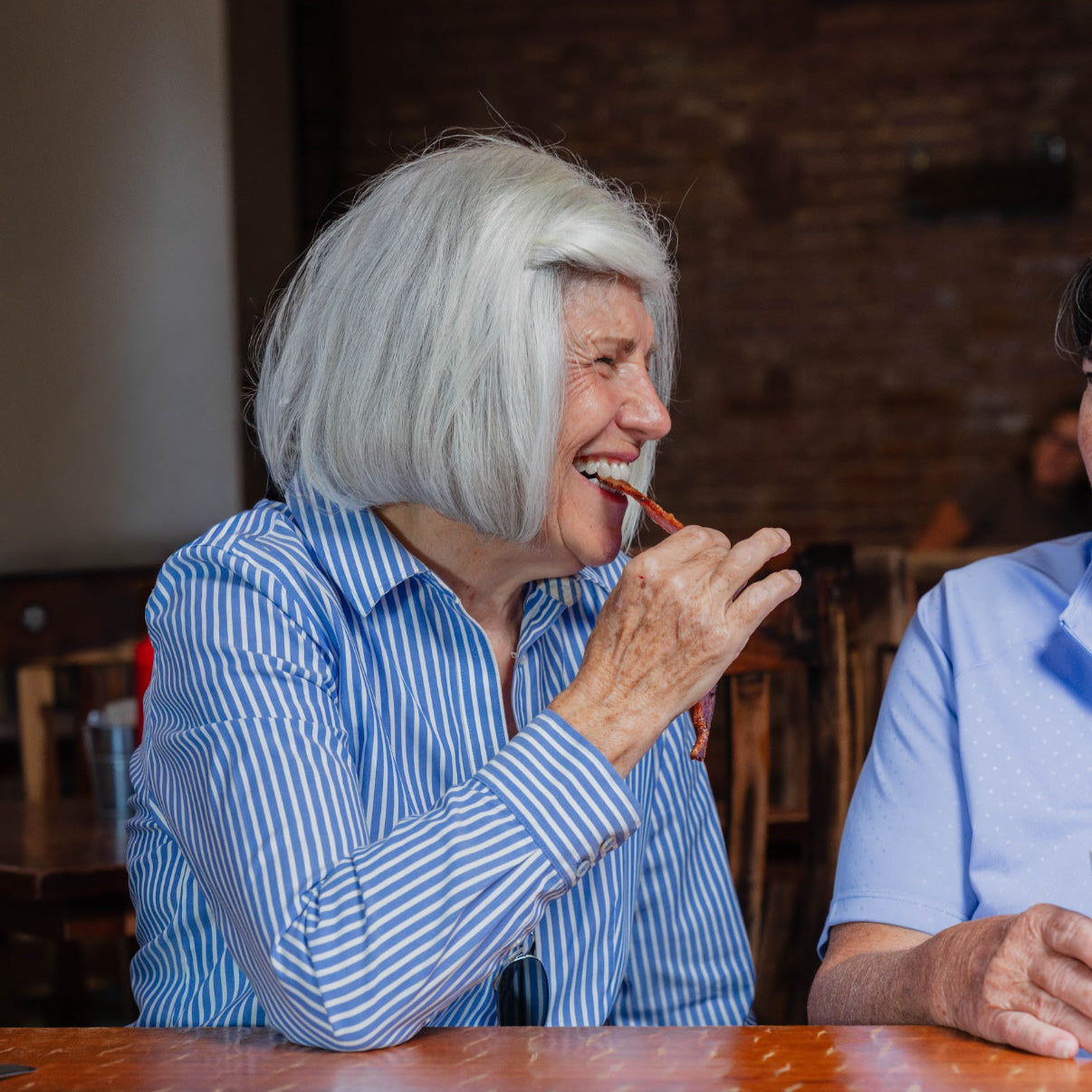 Woman with gray hair eating a bacon strip at a table with another person.