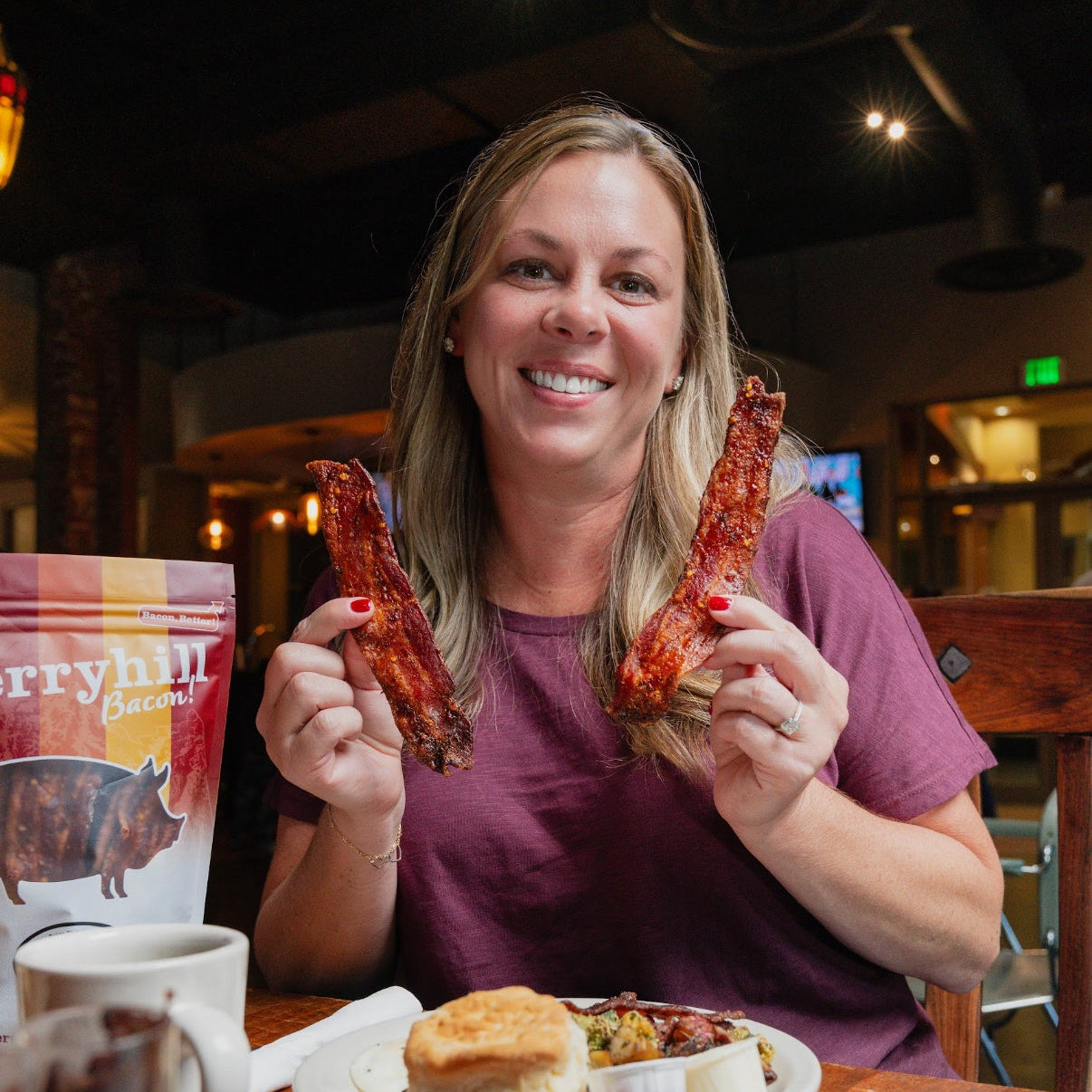 Woman holding two pieces of bacon with a plate of food and a mug on a table in a restaurant.