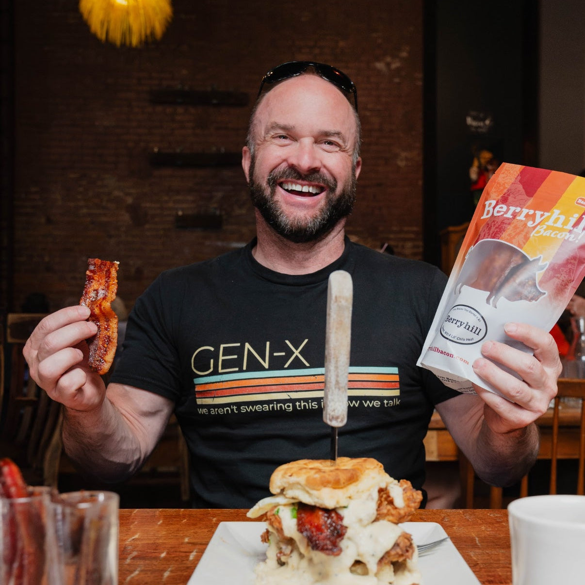 Man holding a sandwich, a package of bacon, and a mug in a casual dining setting.