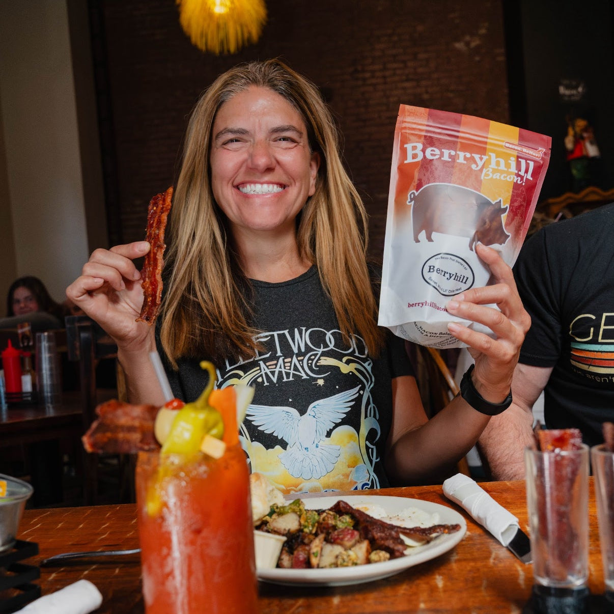 Woman holding a 'Berryhill' product at a dining table with a Bloody Mary and food.