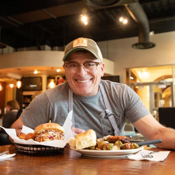 A photo of John Berryhill in a hat and glasses smiling in front of two dishes at BACON Boise restaurant. 