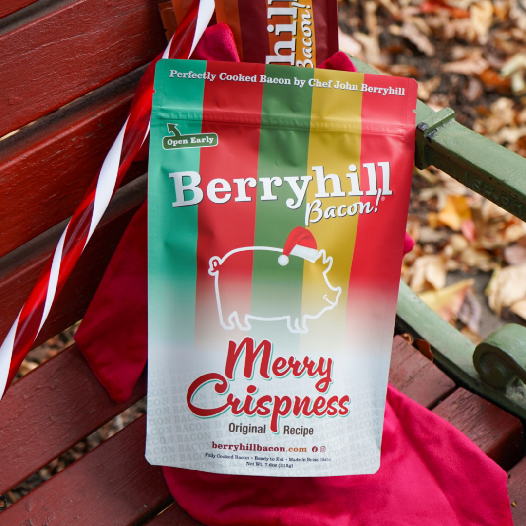 A stand-up pouch of Berryhill Bacon 'Merry Crispness' Original Recipe bacon sits on a red holiday sash and red fabric on a wooden bench outdoors. The packaging features a green-to-red gradient with a white silhouette of a pig wearing a Santa hat, and red and white lettering.