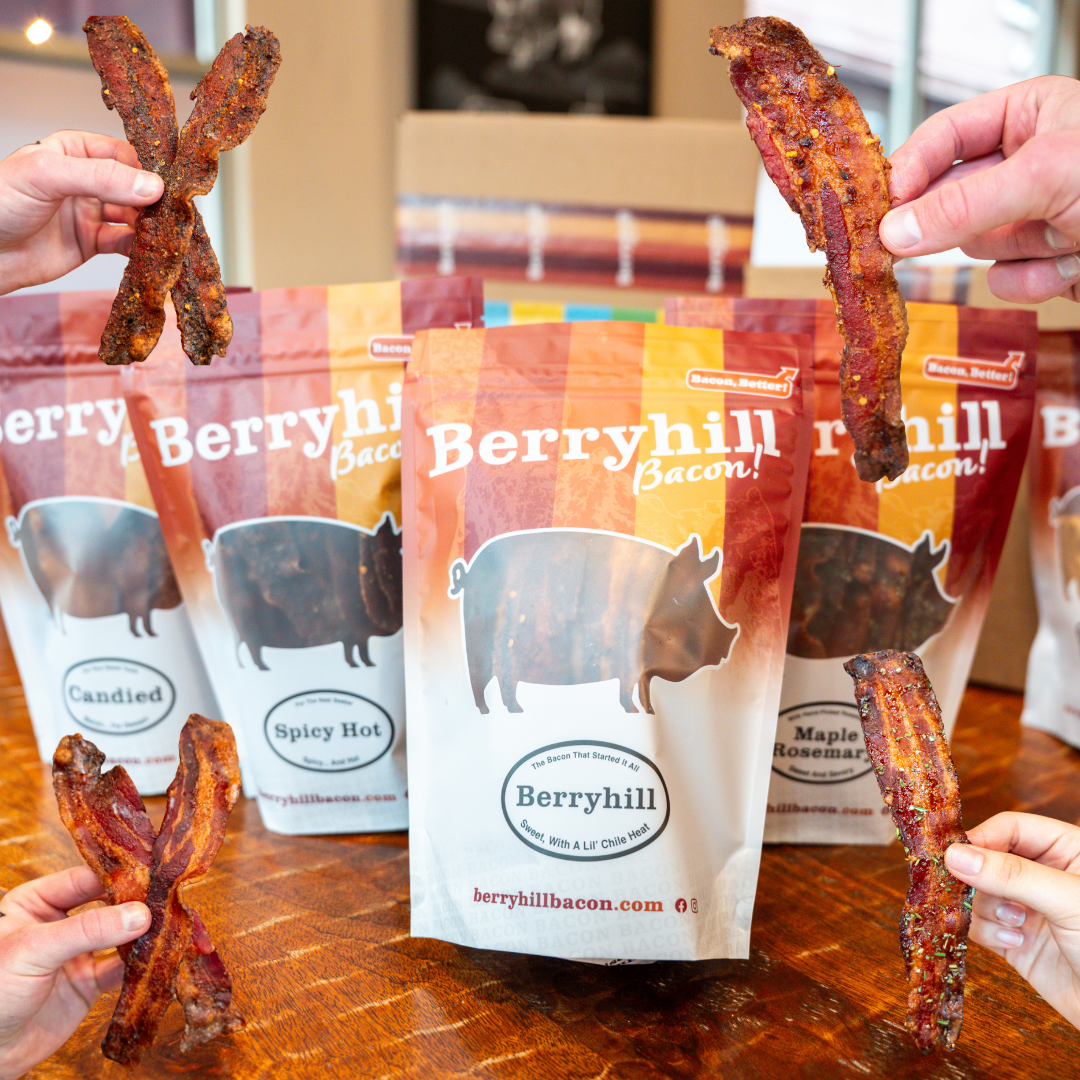 People's hands holding up strips of Berryhill bacon in front of packages with different flavor labels: Berryhill, Spicy Hot, Maple Rosemary, and Candied.